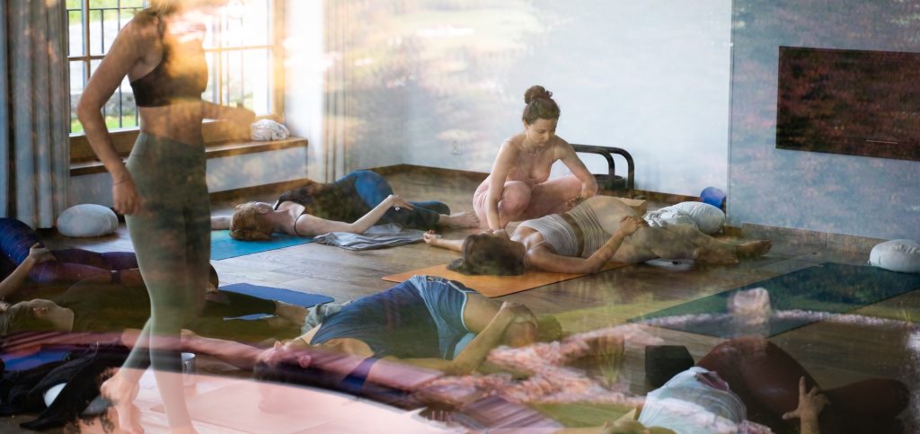 Na obrázku sa nachádzajú ľudia ležiaci za oknom na jogových podložkách v twistoch v lehu. Jeden z ľudí dostáva asistenciu od učiteľa jogy.